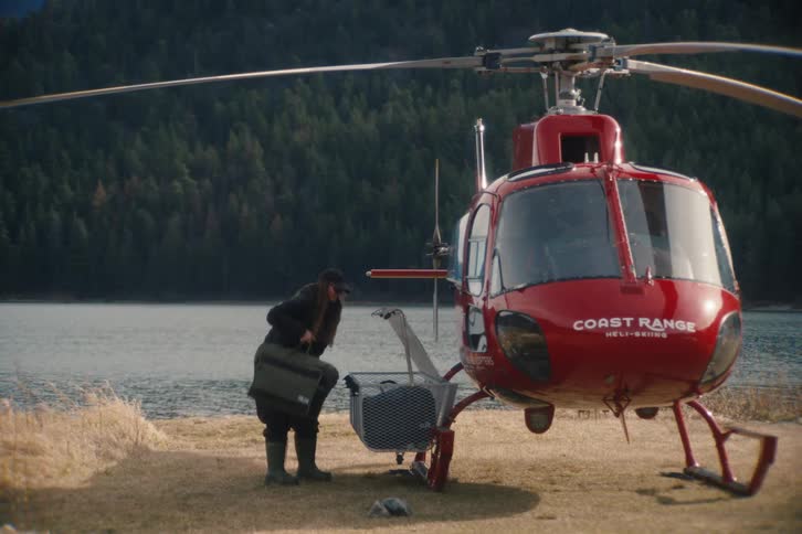 A woman loads her gear and luggage into the cargo basket of a red helicopter parked on the shore of a remote mountain lake