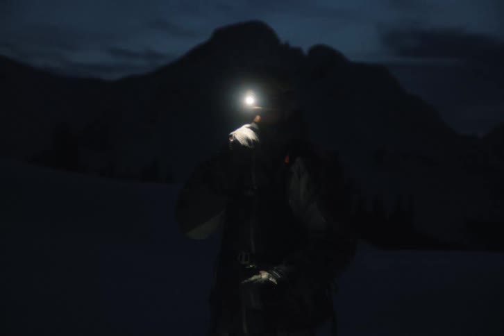 A lone skier with a headlamp walks toward the camera and adjusts their gear in the dark on a snowy mountain