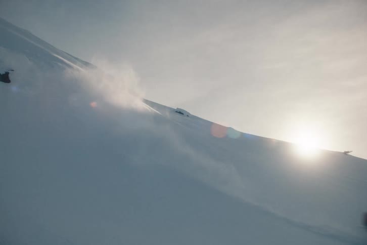 An extreme snowboarder carves down a steep, backlit mountain, kicking up powder snow in slow motion