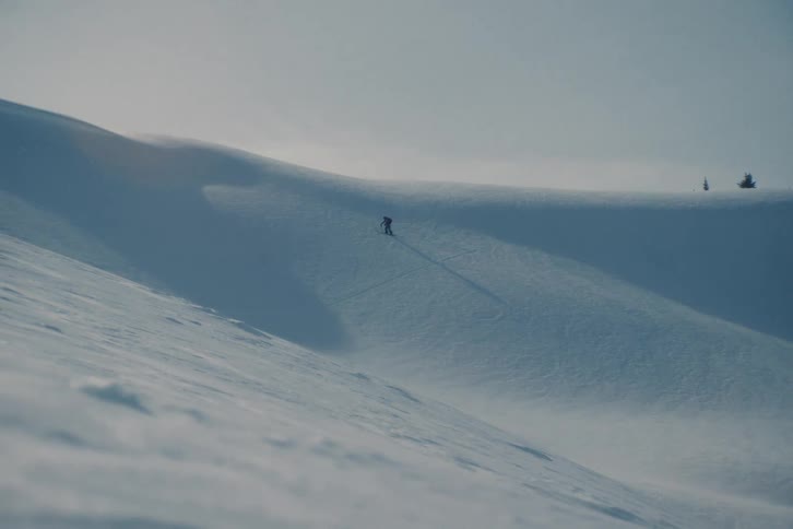 A solitary skier pauses on a vast, pristine snowy mountainside in a minimalist winter landscape