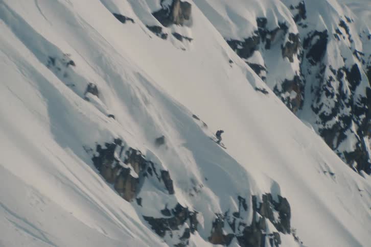 A professional skier carves down a steep, rocky, and snow-covered mountain face before jumping off a cliff in the backcountry