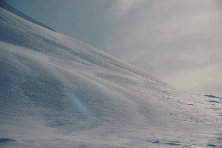 Strong winds blow fresh powder snow across a desolate, sunlit mountainside during a blizzard