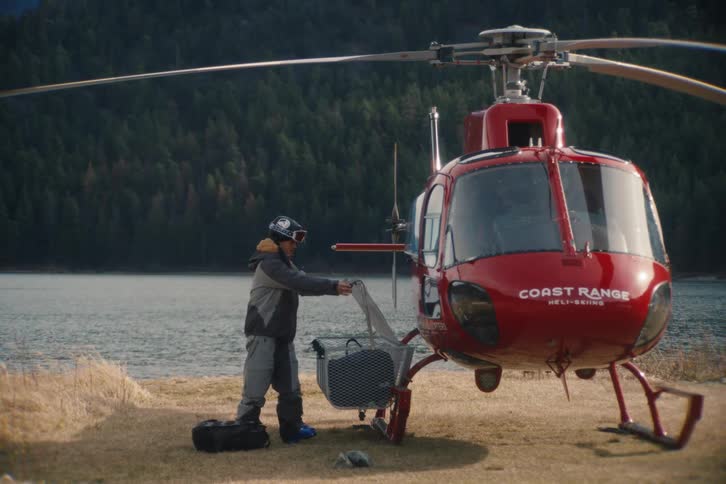 A snowboarder loads their luggage into the cargo basket of a red helicopter parked on the shore of a mountain lake before a heli-skiing trip