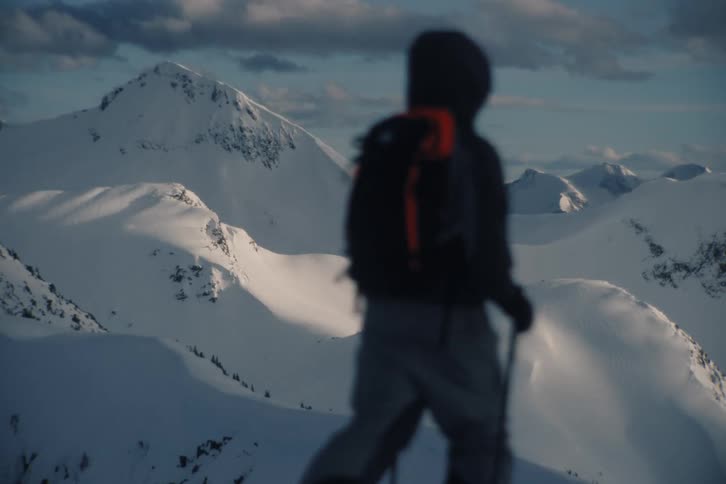 A lone hiker with a backpack and poles walks in front of a stunning, snow-covered mountain range at sunrise