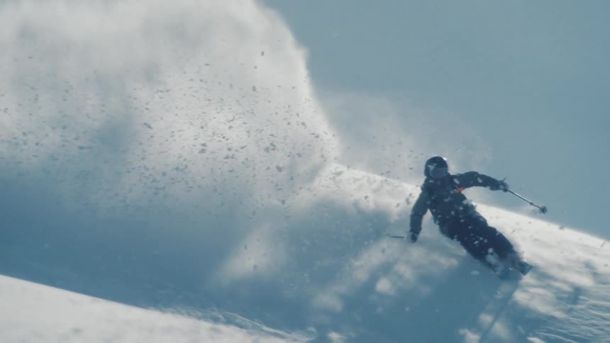 An expert skier carves through deep powder snow down a pristine mountain slope, kicking up a massive spray of snow in super slow motion