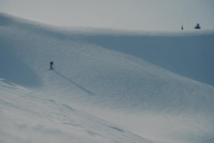 A lone skier with a backpack treks across a vast, windswept snowscape in the remote backcountry