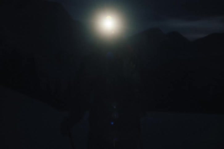 A skier wearing a bright headlamp stands alone in a dark, snowy mountain landscape as the camera slowly zooms in for a close-up