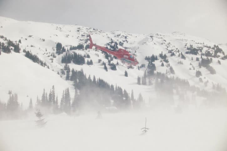 A red helicopter takes off from a remote snowy mountain camp, creating a blizzard of snow as it flies away