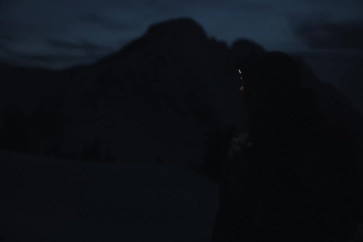 An adventurer walks across a snowy mountain landscape at night, guided by the bright light of a headlamp creating a lens flare