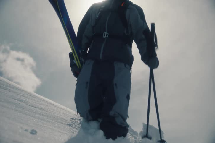 A skier hikes up a snowy mountain in slow motion, silhouetted against a bright sun and a beautiful halo