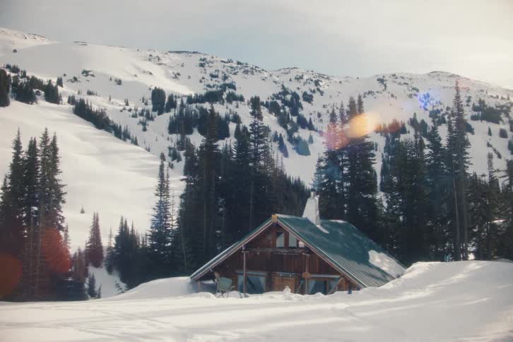 A cozy wooden cabin sits nestled in deep snow at the base of a vast mountain on a bright winter day