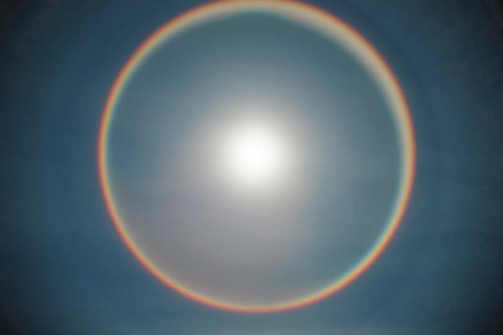 A bright sun with a lens flare effect creates a stunning circular rainbow halo in the sky over a snowy mountain landscape
