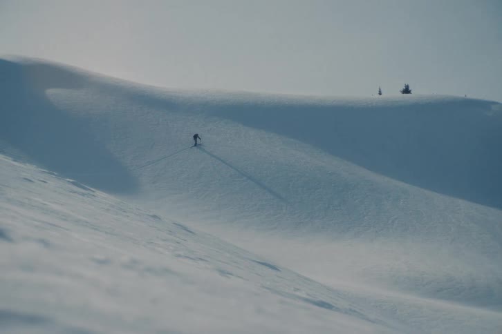 A lone backcountry skier hikes up a vast, windswept, snow-covered mountain, emphasizing the scale of the winter landscape