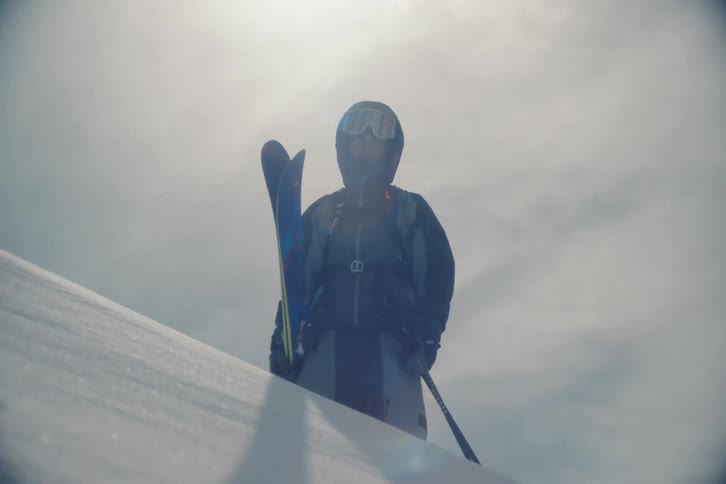 A skier in full gear stands proudly on a snowy mountain top, backlit by the bright, hazy sun