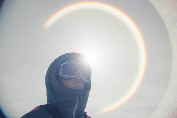A cinematic low angle shot shows a man in a hooded winter jacket and ski goggles as a circular rainbow lens flare from the sun slowly overexposes the shot