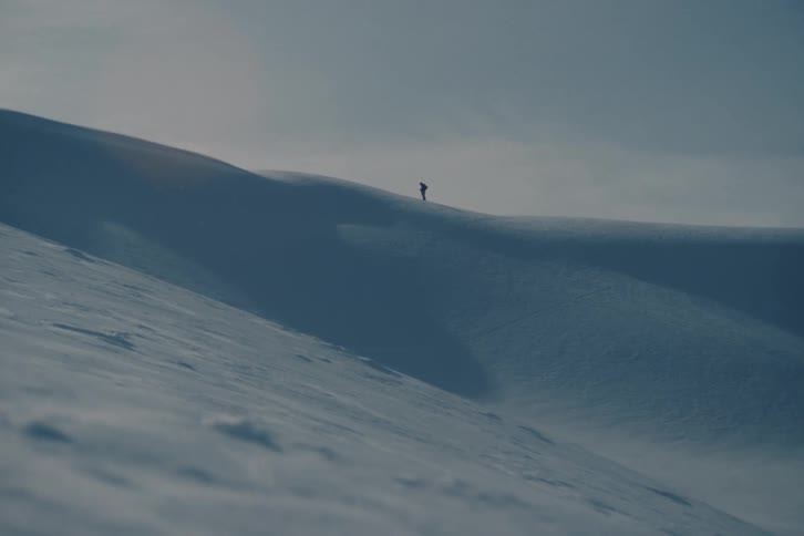 A distant silhouette of a hiker treks across a vast, windy, and snow-covered mountain ridge in a minimalist landscape