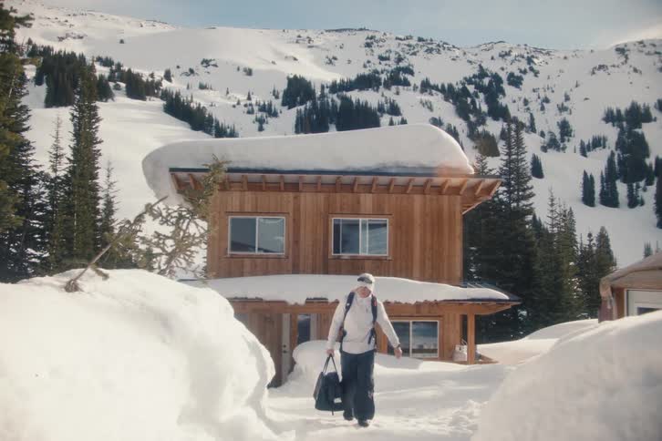 A man in full winter gear leaves a remote wooden ski cabin in the mountains on a sunny day