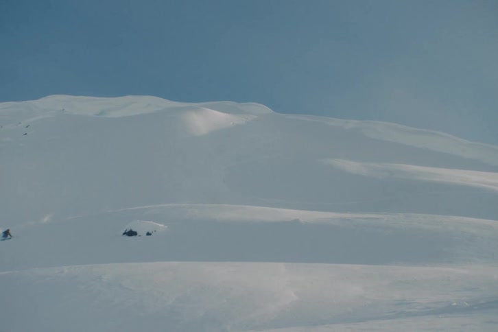 A lone skier carves fresh powder turns down a vast, untouched mountain face in slow motion