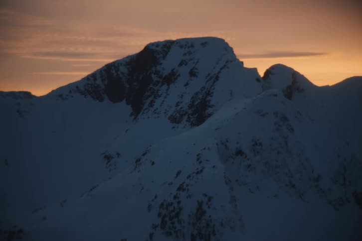 A majestic snow-capped mountain peak stands silhouetted against the warm glow of a winter sunset