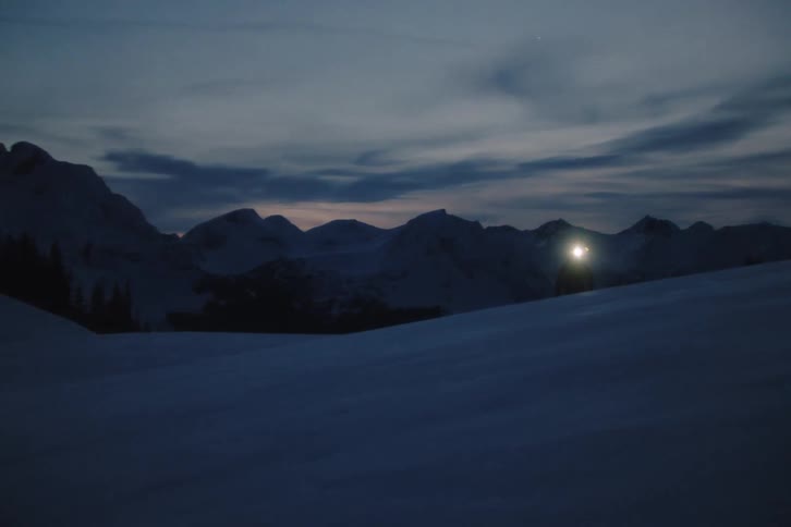 A solitary touring skier with a bright headlamp hikes up a snowy slope towards the camera in the dark of night