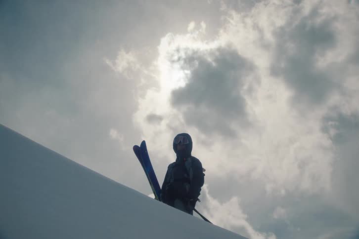A silhouetted skier in full gear stands atop a snowy mountain with their skis, looking out from under a dramatic, sun-drenched sky