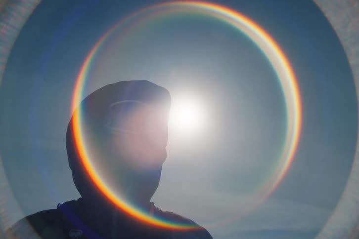 A person in full winter gear turns to face the camera as a beautiful rainbow lens flare forms around the sun