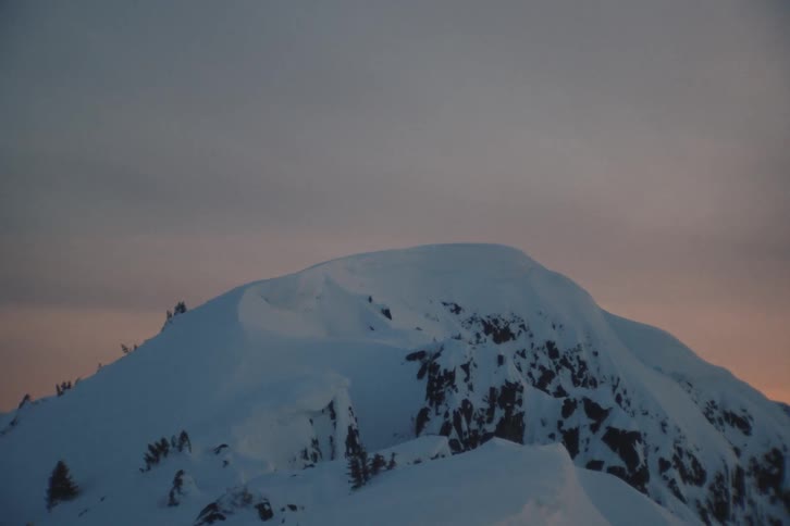 A serene shot zooms in on a snow-capped mountain peak during a beautiful, soft-hued dusk
