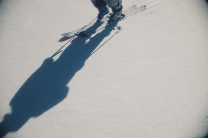 The shadow of a back country skier moves across untouched snow, followed by the skier's legs and skis, leaving fresh tracks