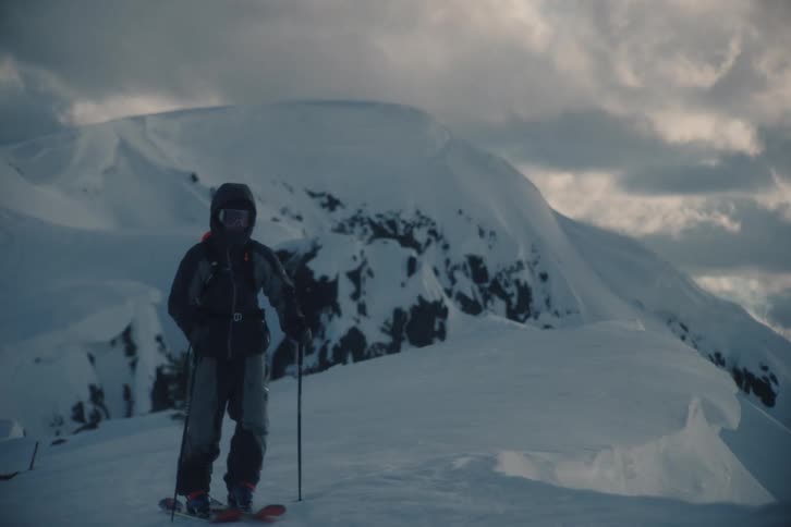 A lone skier slowly hikes towards the camera along a snowy mountain ridge under a dramatic, cloudy sky