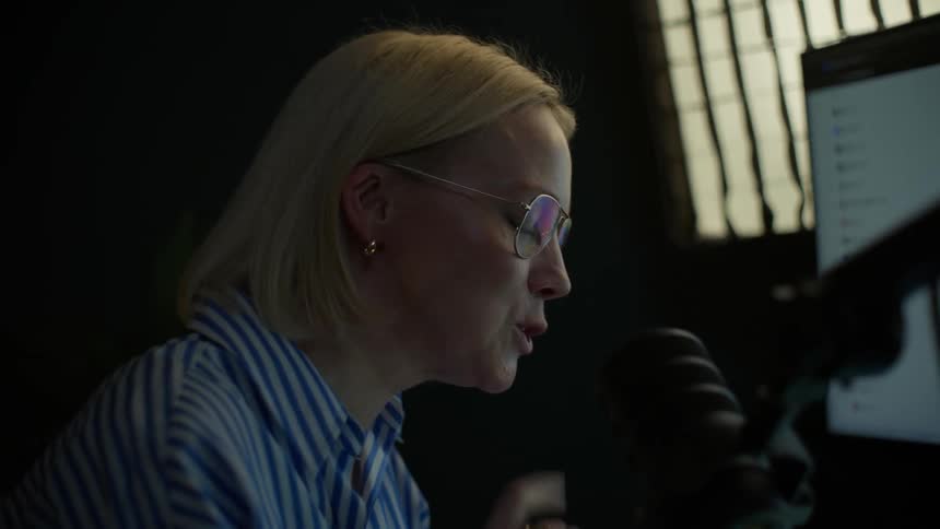 A female podcaster with glasses speaks animatedly into a microphone while working on her laptop in a dark studio