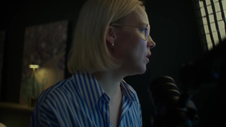 A professional female podcaster with glasses records an episode in her dimly lit home studio, speaking into a microphone