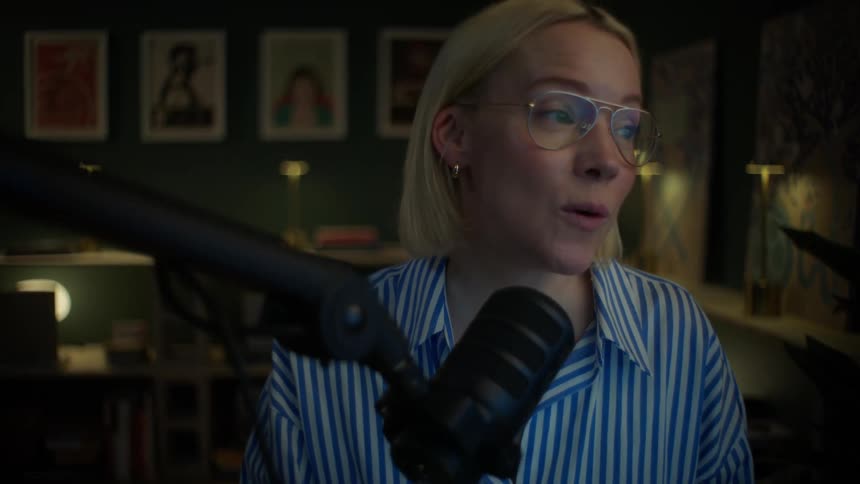 An expressive podcaster with short blonde hair laughs and talks into a studio microphone during a recording session