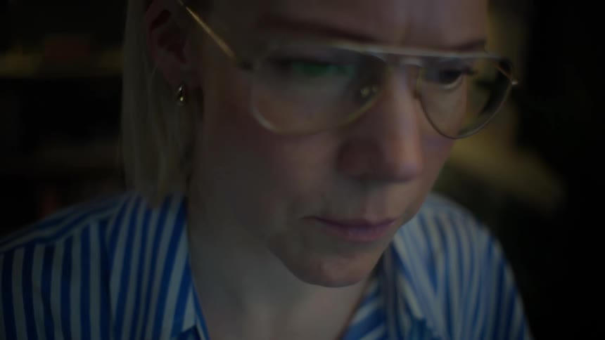 A close-up shot of a focused woman with glasses working late at night on her laptop, the screen illuminating her face