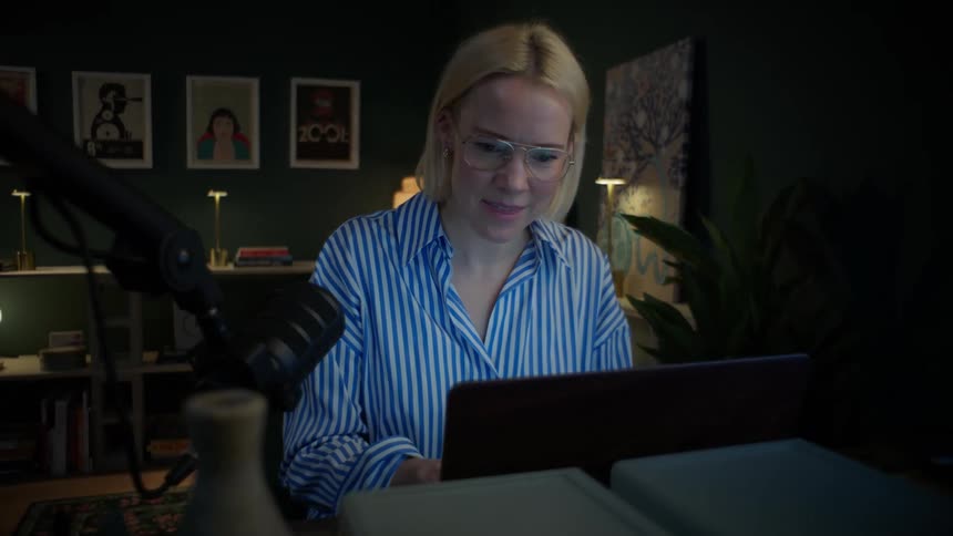 A female content creator with glasses works at her desk at night, her expression changing to serious as she reads her screen