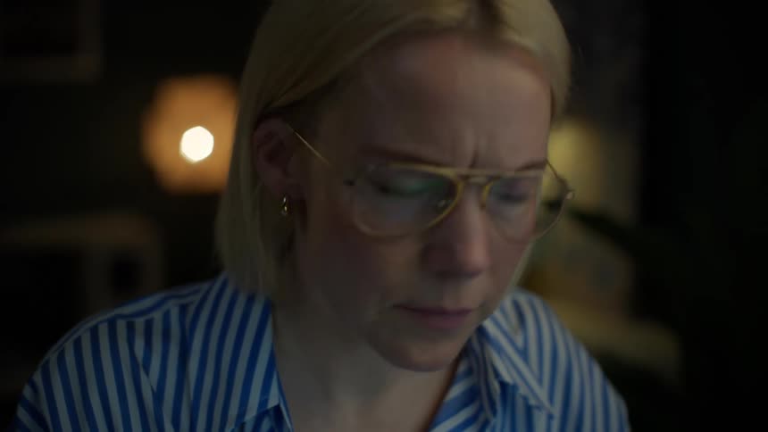A serious woman with blonde hair puts on her glasses and focuses intently while working in a dimly lit room