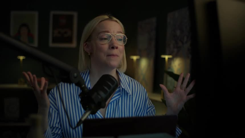 A young woman with blonde hair and glasses enthusiastically records audio for a podcast in a stylish, dimly lit home studio