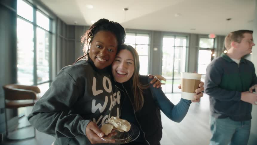 Two happy diverse female friends embrace and smile for the camera while enjoying coffee and breakfast in a sunlit community lounge
