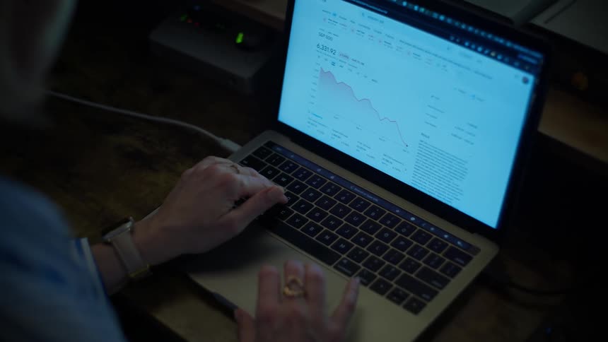 An over-the-shoulder view of a woman analyzing a declining stock market chart on her laptop in a dark room