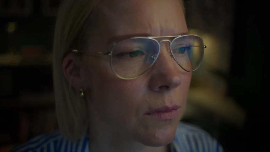 A focused woman with glasses works on her laptop in a dimly lit home office, her face illuminated by the screen as the camera slowly zooms in