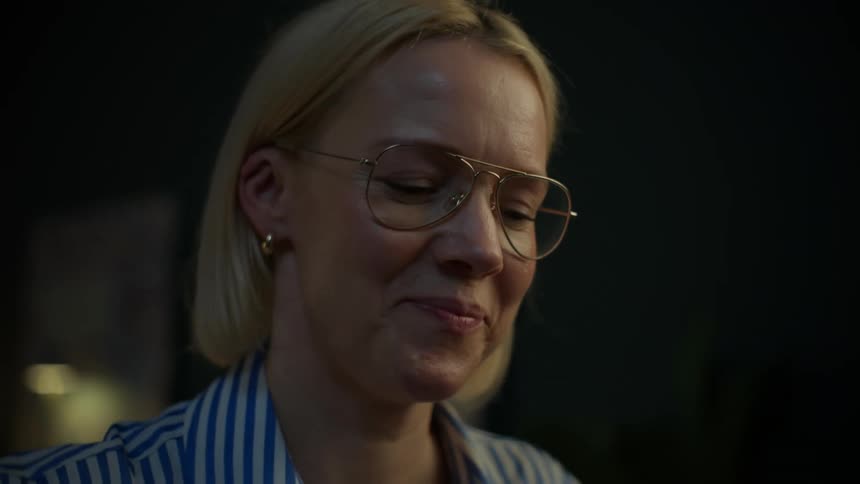 Close-up of a smiling blonde woman with glasses who is happily working late on her laptop in a dimly lit office