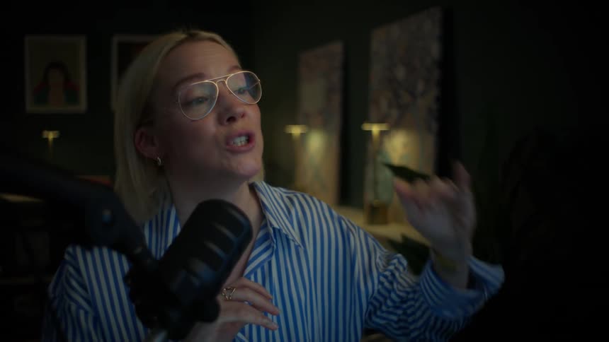 A blonde woman with glasses enthusiastically speaks into a microphone while recording a podcast in her stylish home studio at night