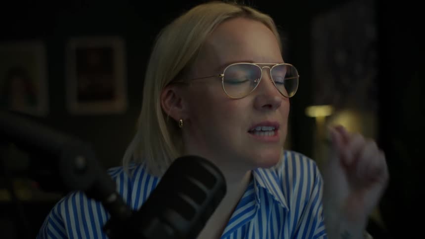 Close-up slow motion shot of a woman with glasses speaking passionately into a microphone while working on her laptop in a dark home studio