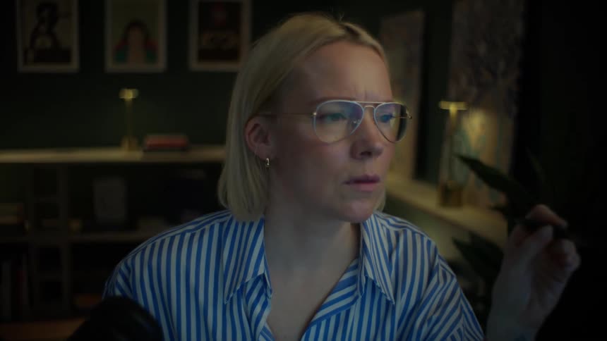 A focused woman with glasses works late on her computer in a dimly lit home office, concentrating intently on her screen