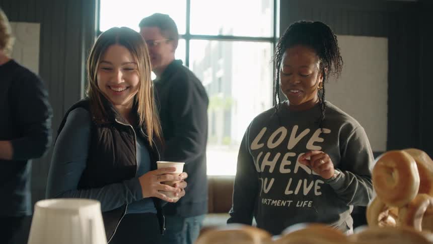 Two diverse women enjoy coffee and bagels while socializing in a modern apartment community lounge