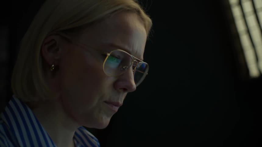 A dedicated professional woman works intently on her laptop, her face illuminated by the screen in a dark room