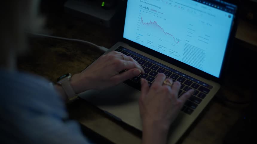 A businesswoman works late at her desk, typing on her laptop while tracking a falling stock market chart