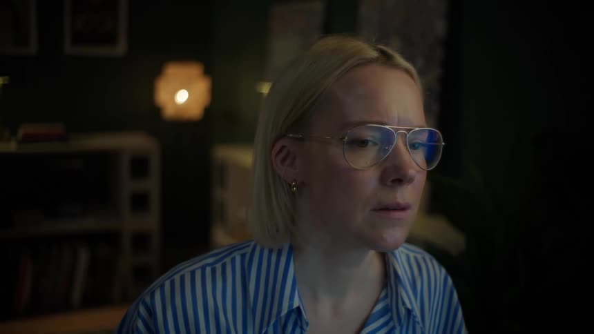 A focused blonde woman wearing glasses stares at her screen with a concerned expression while working from her home studio at night