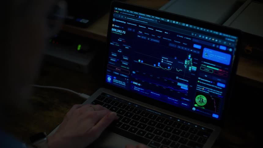 Over-the-shoulder shot of a person monitoring the fluctuating Bitcoin market on a laptop screen in a dark room