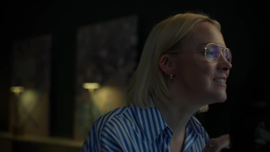 A blonde woman wearing glasses speaks animatedly and yawns during a late-night conversation in a dimly lit room