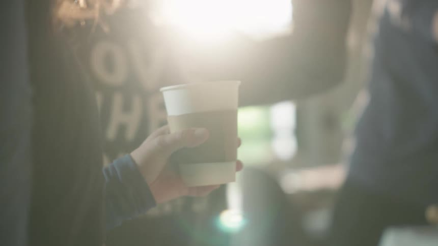 A person holds a steaming hot cup of coffee, gesturing while talking with others in a brightly lit, social setting in slow motion